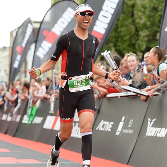 Domenico Buonocore running down the finish chute wearing a black and red tri suit grimacing with his arms outstretched by his side